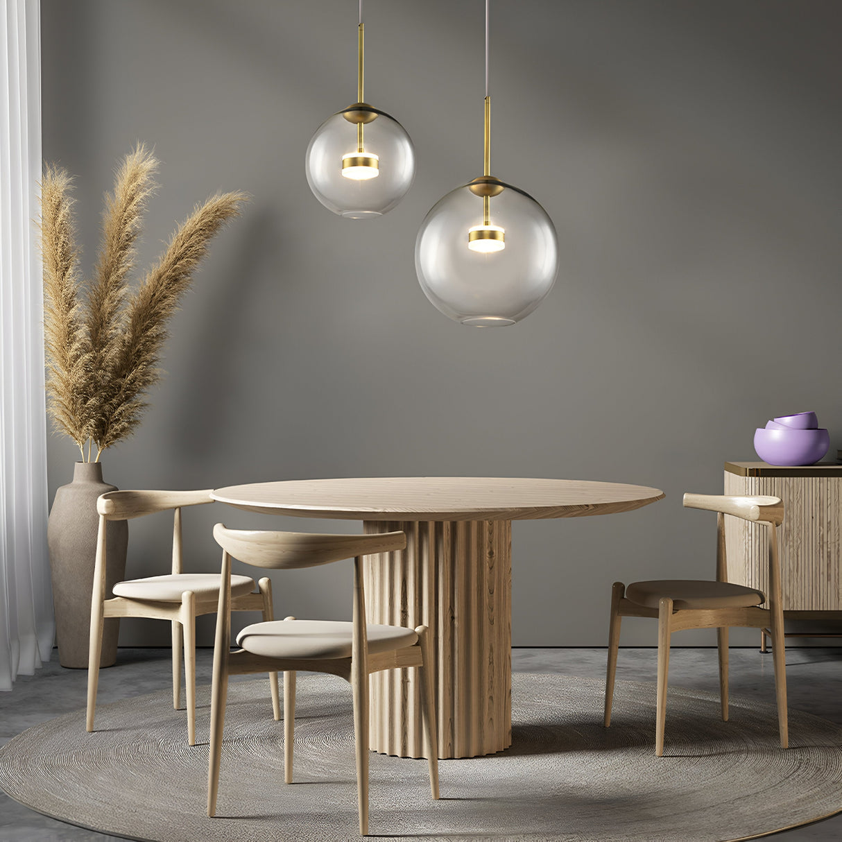 A minimalist dining room boasts a round wooden table with three matching chairs on a textured rug. Two Cometa LED Pendant Lights in brushed gold illuminate the space warmly. Pampas grass fills a corner vase, and a small cabinet nearby holds a purple bowl.
