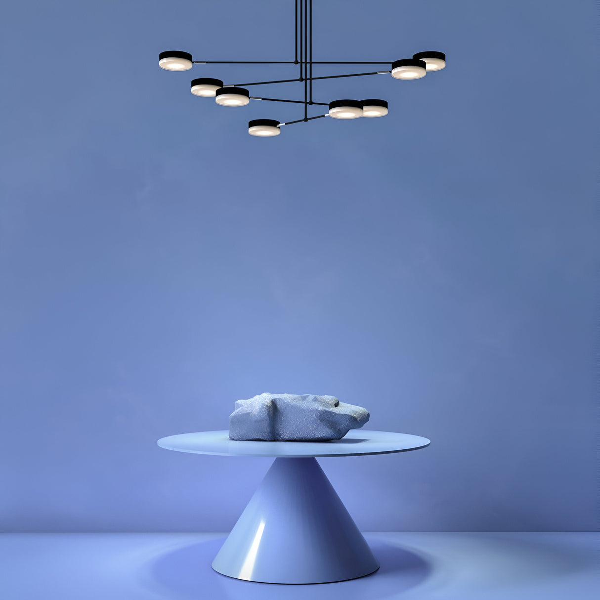A modern minimalist scene features the Fad LED 8 Light Chandelier - Black with LED, geometric design illuminating above a round pedestal table. On the table rests an abstract, textured white sculpture set against a sleek blue background.