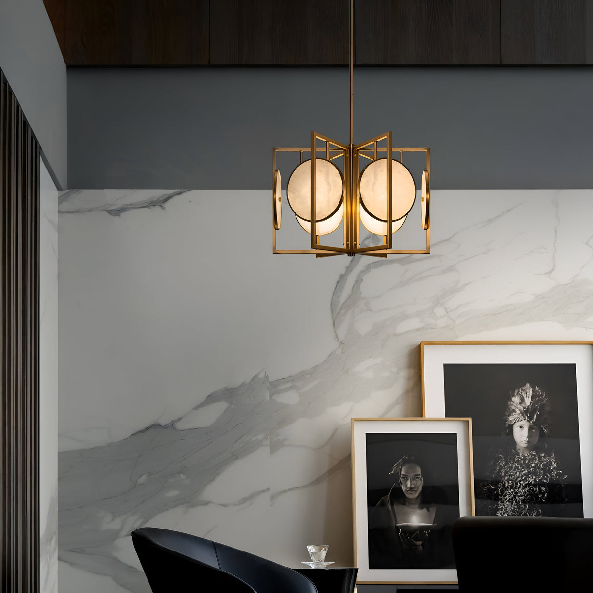 A modern interior showcases the Marmo 6 Light Chandelier in gold with stone plates, a marble-patterned wall, and framed black-and-white portraits against a gold-plated frame. A black chair and a small table with a cup complete the scene.