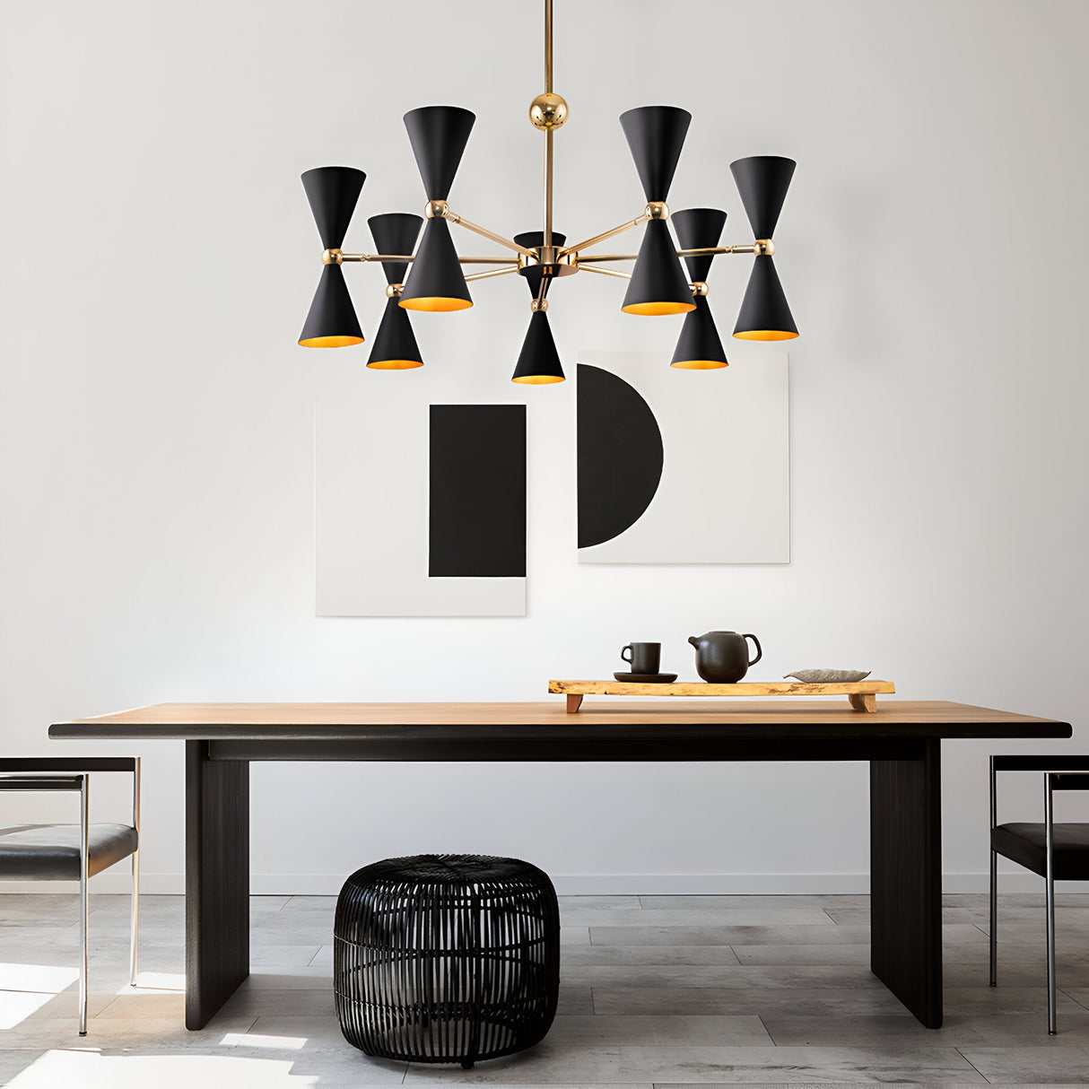 A minimalist dining room with a black table, wooden surface, and two benches features the Vesper 14 Chandelier in gold and black for elegance. Two abstract black and white artworks hang on the wall, with a small black woven ottoman beneath the table.