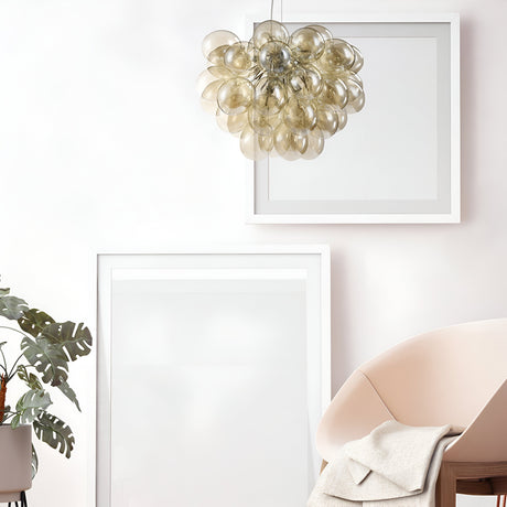 A modern interior includes a round beige chair with a light blanket, two large white-framed picture frames, a green plant, and the Balbo 8 Light Chandelier - Nickel with Cognac Glass. This pendant light features clustered orbs against a light wall.