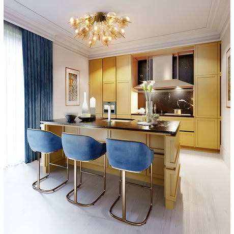 A modern kitchen showcases yellow cabinets and a black marble island, lined with three blue velvet bar stools. Above, the Bolla 4 Light Semi Flush in brass with amber glass hangs. The space is completed by a large window with blue curtains, wall art, and stainless steel appliances.