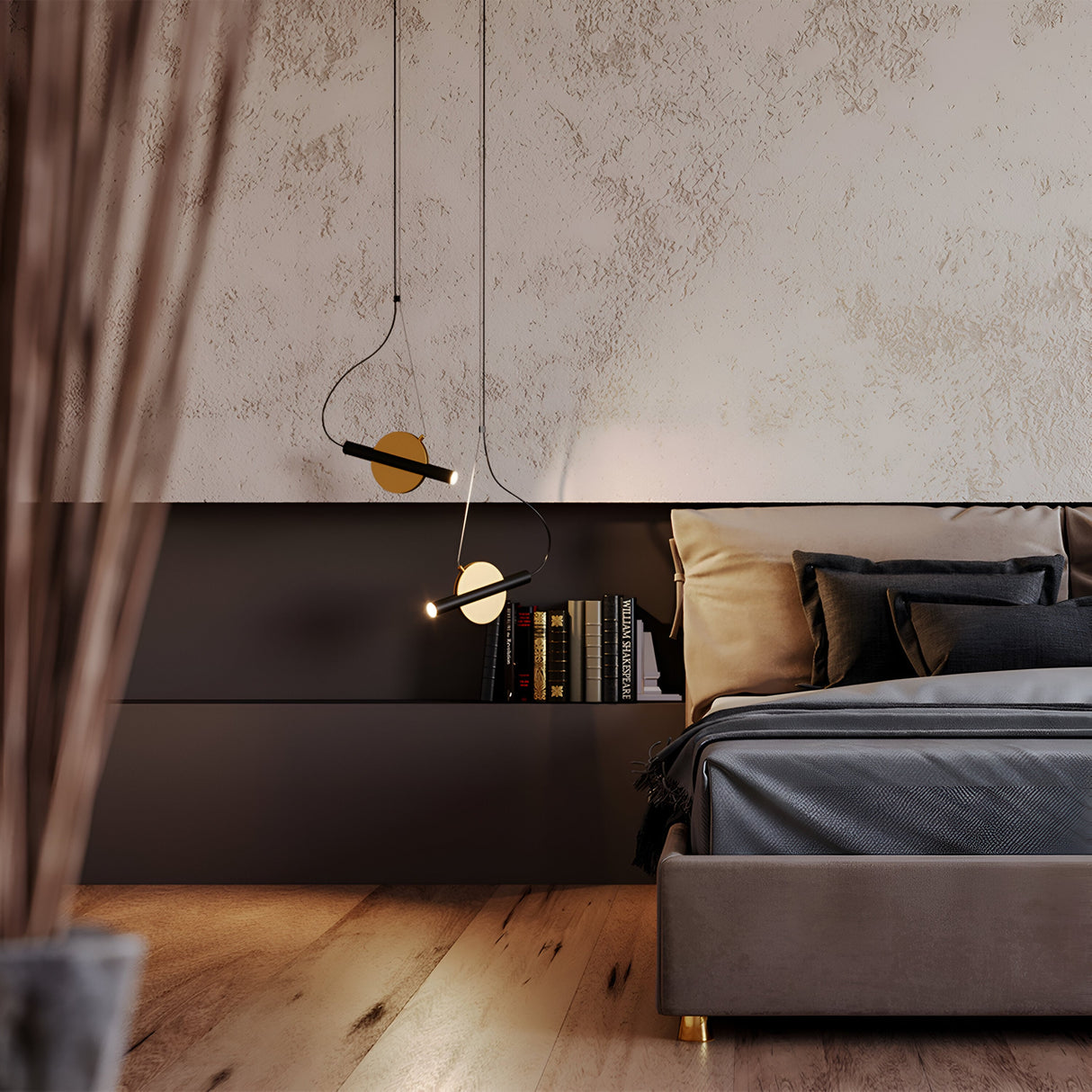 A modern bedroom with a textured beige wall and dark paneling features the Enigma Adjustable LED Pendant Light in brass with black above a shelf of books. The bed, decorated with black and beige pillows, accents the warm-toned wooden floor while blurred sticks add depth in the foreground.