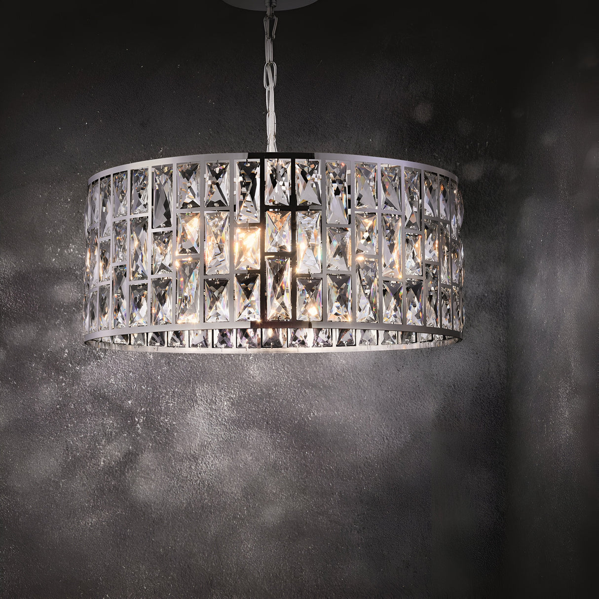 The Gelid 4 Light Chandelier in chrome features a round design with geometric crystal glass patterns, casting a soft light on a textured dark gray wall.