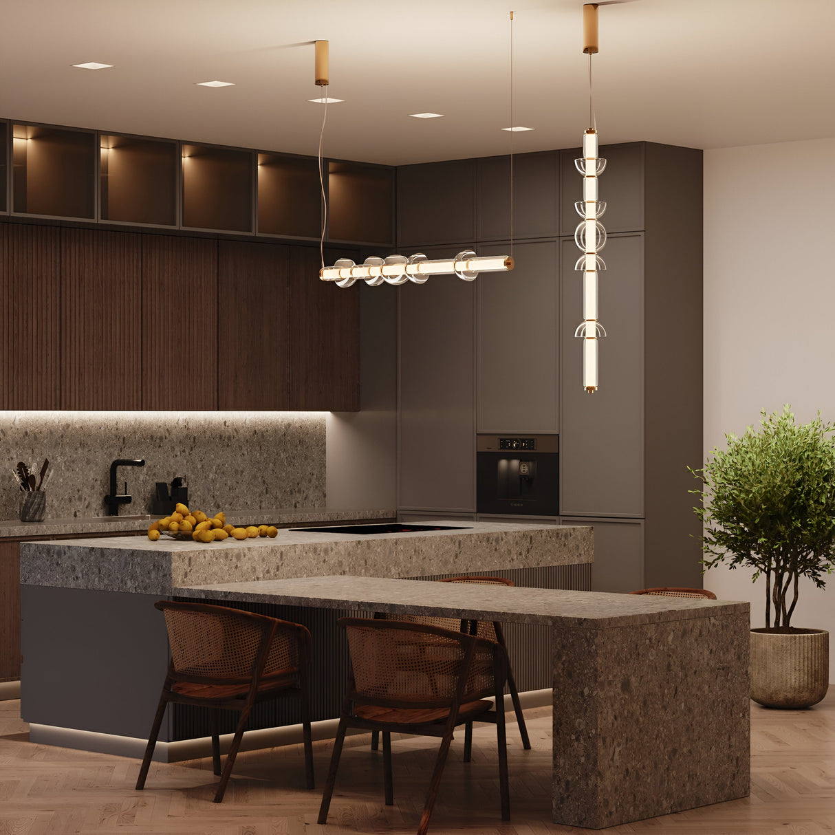 A modern kitchen features a large island with a stone countertop. The Cosmo LED Linear Suspension Light in brass with clear glass hangs from the ceiling, illuminating the space. Dark cabinets enhance the sleek design, while a bowl of lemons and a potted plant add color and greenery.