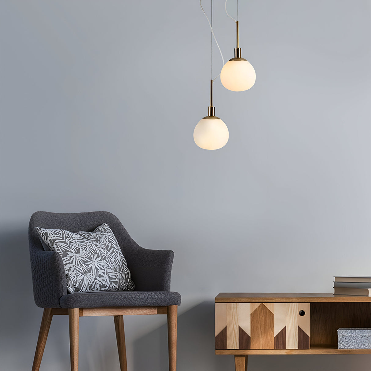A modern interior showcases a dark gray upholstered chair with a patterned cushion, a wooden sideboard with geometric designs, and two Erich 1 Light Globe Pendants in brass with white glass. Their soft lighting enhances the minimalist elegance of the room.
