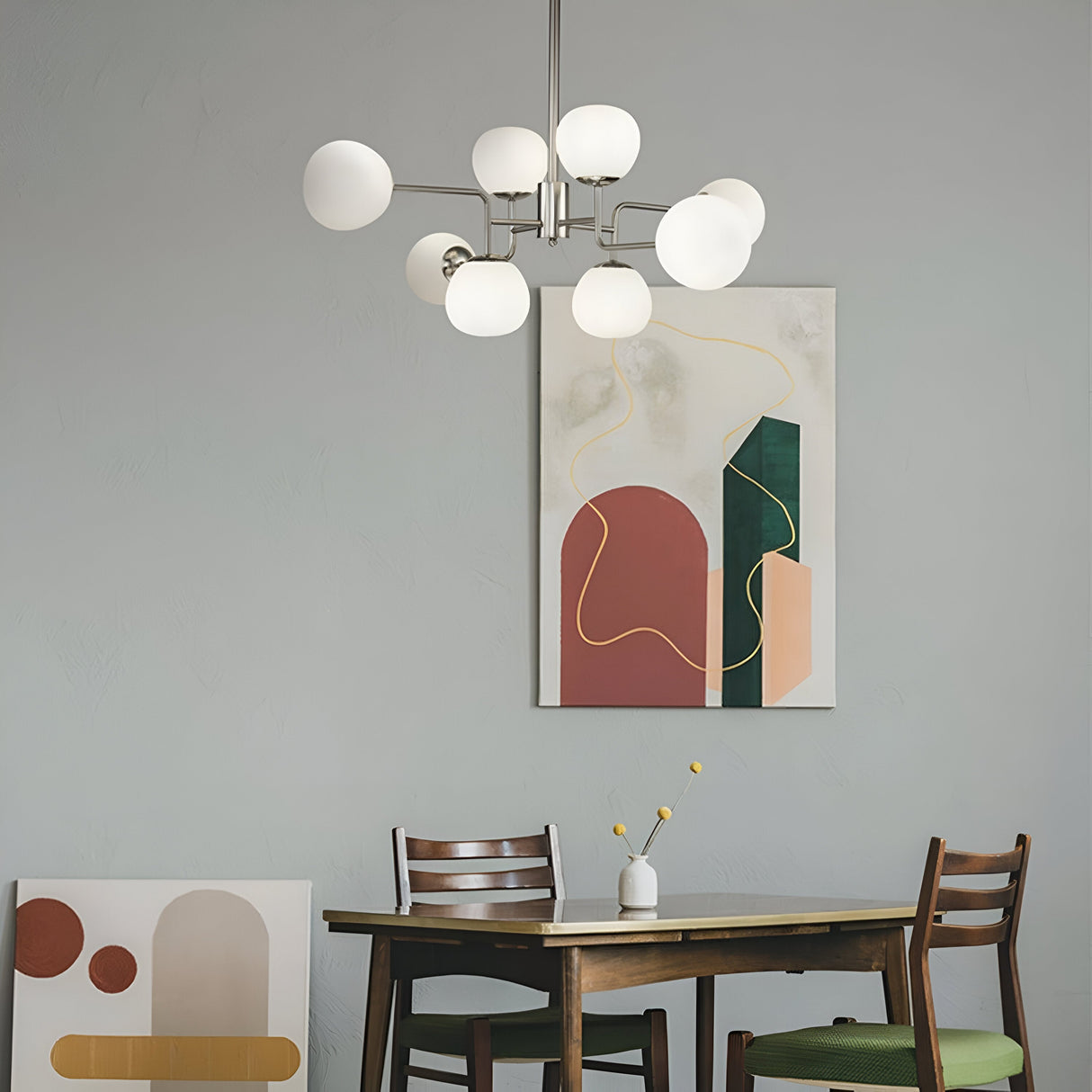 An 8-light Erich chandelier in nickel with white glass illuminates a dining area. Below, a wooden table with a white vase of yellow ball flowers is flanked by two chairs. Abstract art featuring earthy tones and geometric shapes adorns the wall and floor.