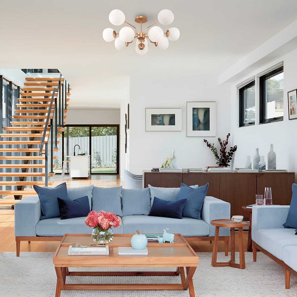 A modern living room features light blue sofas with dark blue cushions and a wooden coffee table adorned with pink flowers and a teapot. Overhead, the chic Erich 10 Light Semi-Flush - Brass With White Glass chandelier presides. A wooden staircase leads upstairs, with the kitchen visible nearby.