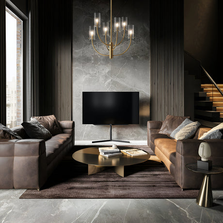 Two brown leather sofas face each other in a sophisticated living room. A large modern TV is mounted on a dark marble wall. A round coffee table with books stands at the center on a plush rug, while an elegant Arco 6 Light Chandelier - Brass with ribbed glass lampshades completes the decor.