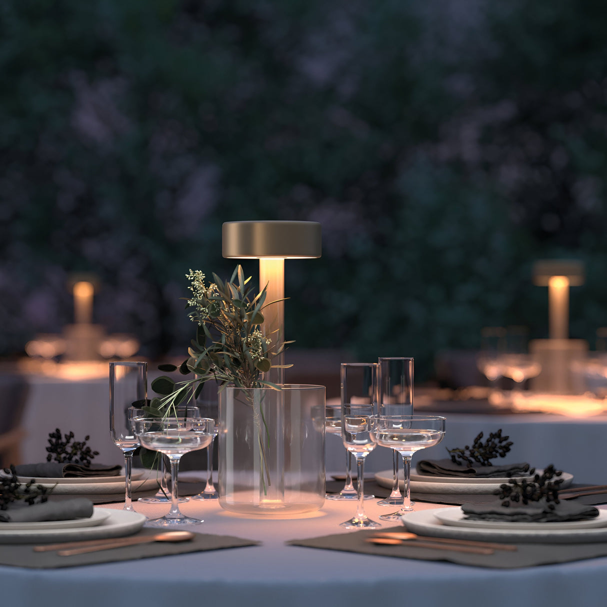 An elegant outdoor dining area showcases the AI Collaboration Rechargeable LED Table Lamp with Glass Vase - Gold, adorned with greenery. Neatly arranged glassware and plates enhance the setting, while a blurred greenery backdrop adds to the serene ambiance.