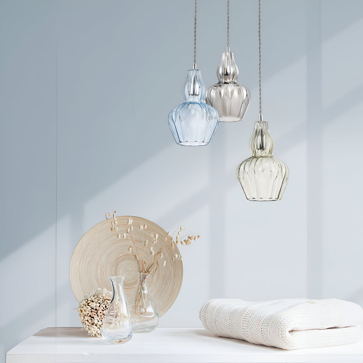 A serene interior space features a Nickel Eustoma Pendant Light with light blue glass, accompanied by others in silver and clear. Below, a white table displays vases, dried flowers, a round wooden decor piece, and a neatly folded white blanket.