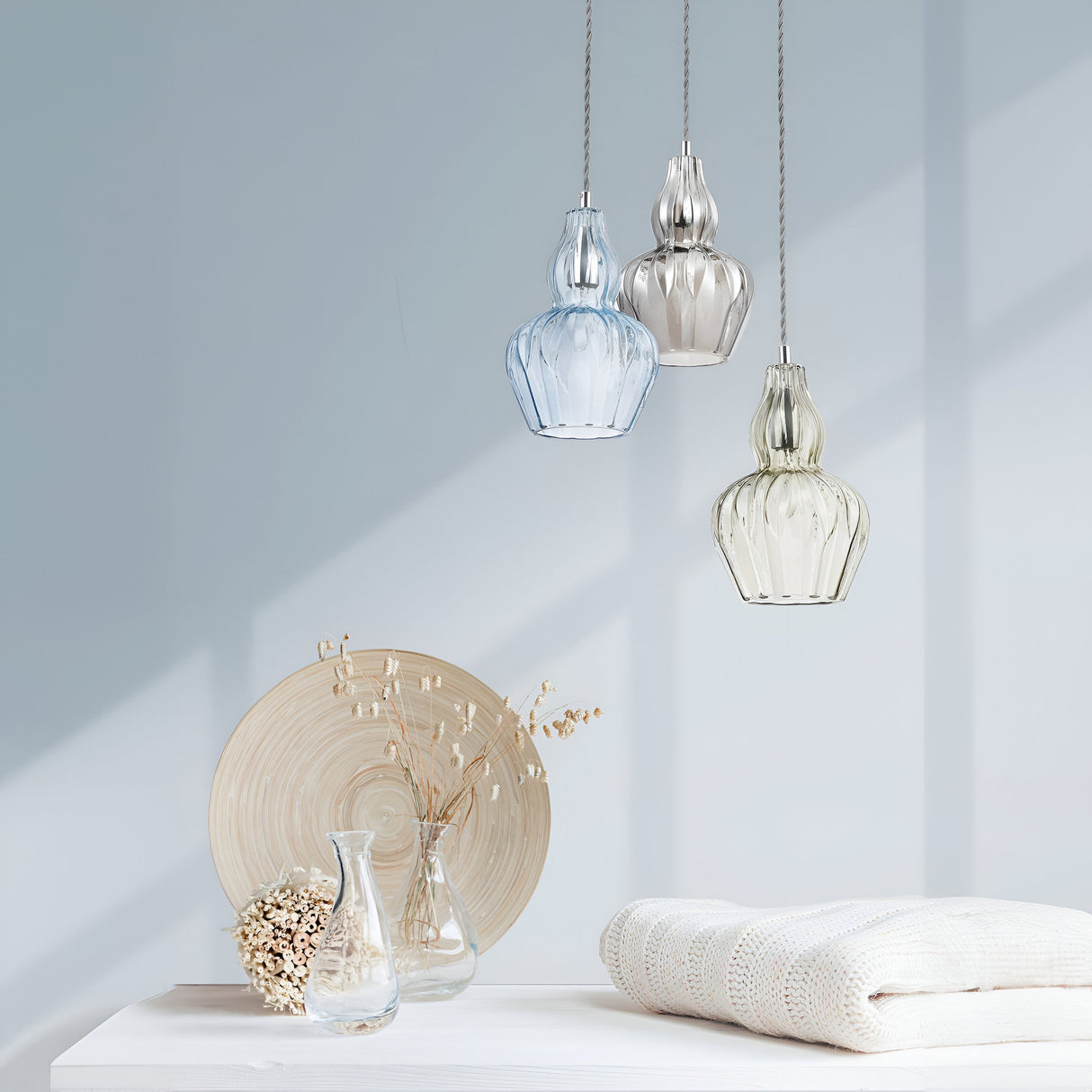 Three Eustoma Pendant Lights - Nickel With Transparent Glass illuminate a table adorned with a wooden plate, two glass vases, and white folded fabric. Natural light enhances the setting, casting gentle shadows on the pale blue background wall.