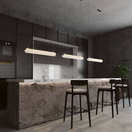 A modern kitchen with a minimalist design showcases a textured stone island, three high chairs, sleek dark cabinetry, and two Petra LED Linear Suspension Lights in Brass With Natural Stone. A green plant against gray-toned walls enhances the contemporary ambiance.