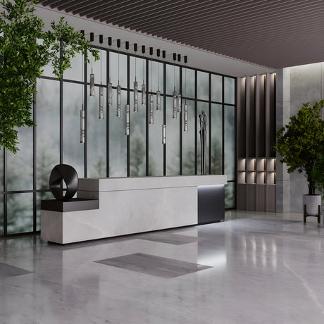 The modern lobby boasts a sleek marble reception desk and Ordo LED Long Pendant Lights with smoky cylindrical glass. Large windows reveal blurred outdoor greenery. A minimalist sculpture adorns the desk, surrounded by artfully placed plants, creating a serene ambiance.