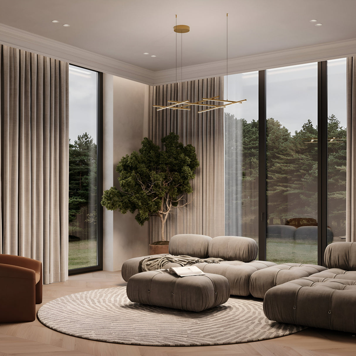 In a modern living room, plush gray sofas and a circular patterned rug are beautifully illuminated by the Radiance LED Suspension Light - Gold. Beige curtains frame floor-to-ceiling windows, while a tree adds nature in the corner, and a geometric satin brass chandelier graces the ceiling.