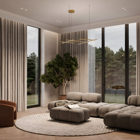 In a modern living room, plush gray sofas and a circular patterned rug are beautifully illuminated by the Radiance LED Suspension Light - Gold. Beige curtains frame floor-to-ceiling windows, while a tree adds nature in the corner, and a geometric satin brass chandelier graces the ceiling.