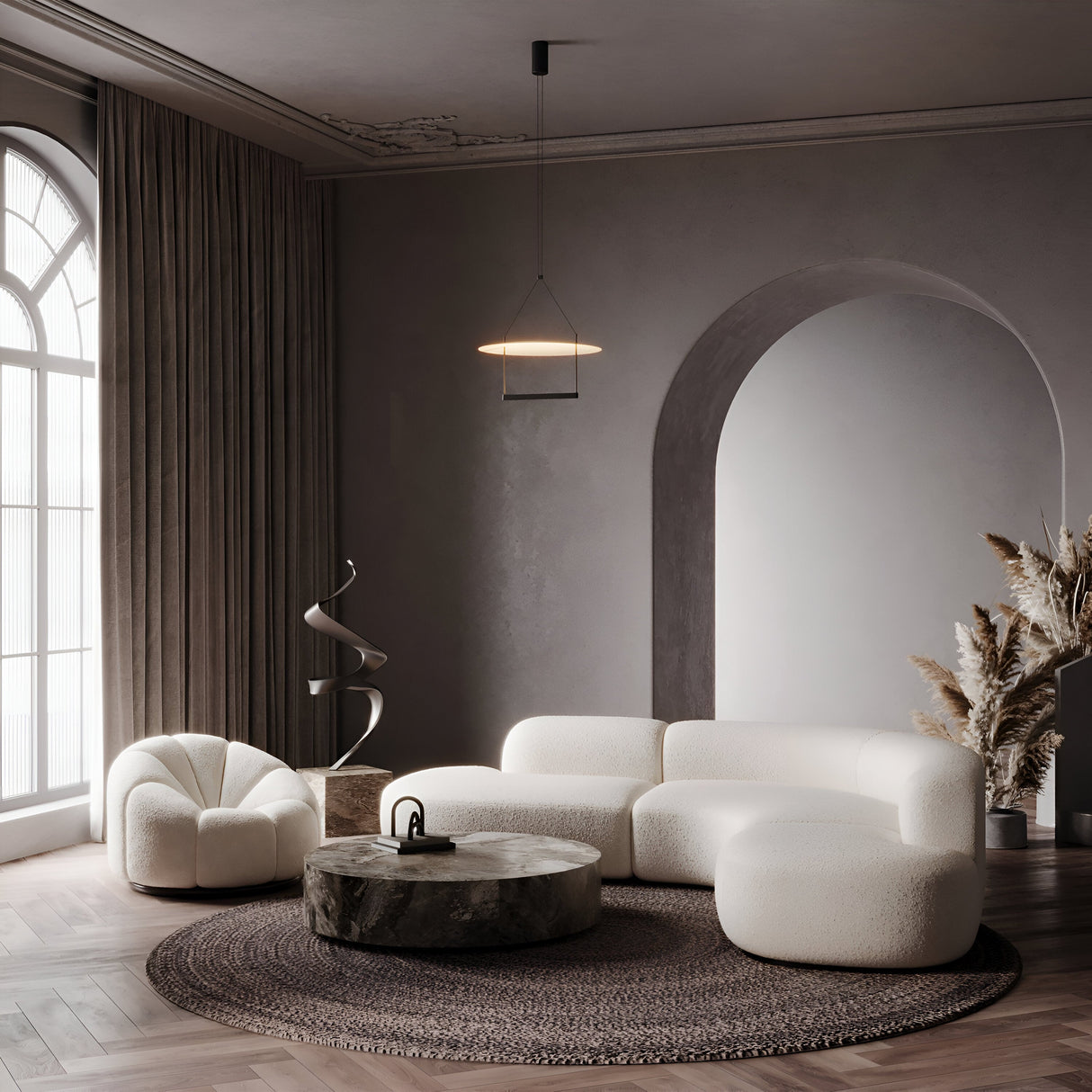 A modern living room embodies minimalist design with a white curved sofa, round ottoman, and textured armchair. A circular wooden coffee table, dried pampas grass decor, floor-length curtains, and a large arched window complement the look. Above it all is the Ambience LED Round Flat Large Pendant Light in black.