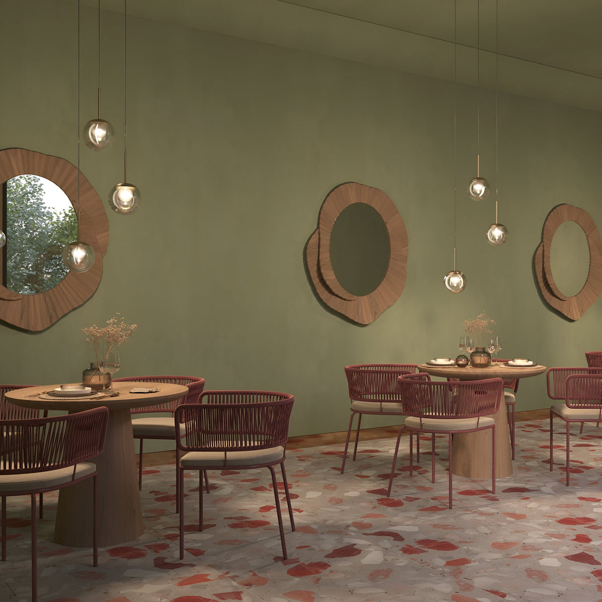 A modern dining area features round wooden tables and red chairs. Circular mirrors decorate the sage green walls, while Basic Form Globe Pendant Lights with gold and frosted/clear glass illuminate from above. The floor showcases a mosaic pattern in shades of red and beige.