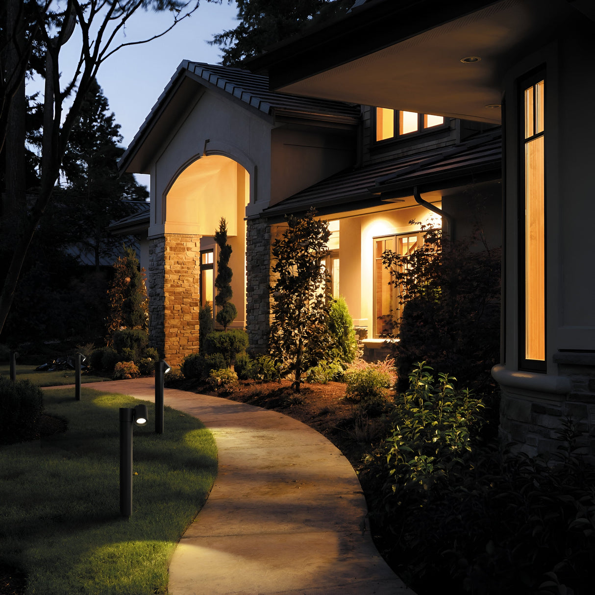 A warmly lit house at dusk, featuring a stone and stucco exterior, showcases a curved concrete path lined with Wall Street 1 Light Bollard 55cm - Black. The path weaves through a garden of shrubs and trees, while large windows glow from within, enhancing the inviting ambiance.