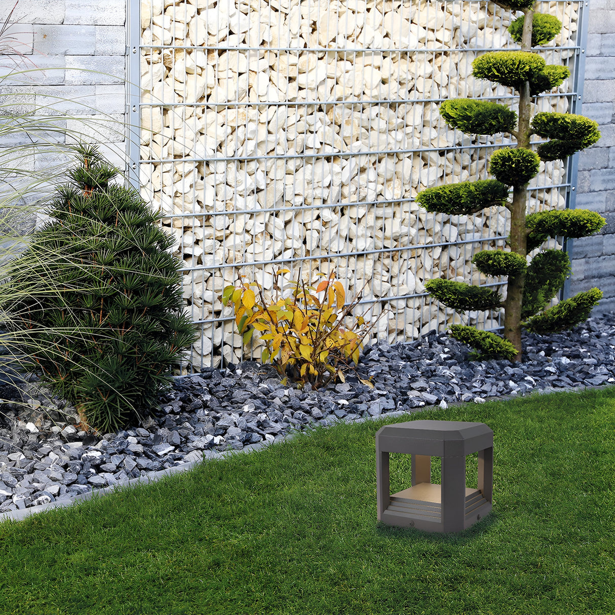 A modern garden features a sculpted evergreen tree, a yellow-leaved bush, and a cylindrical tree. The Royal Mile LED Square Pedestal Light in grey elegantly illuminates the scene, its die-cast aluminum body blending with firewood stacked in the background, as a wooden stool sits on the grass.