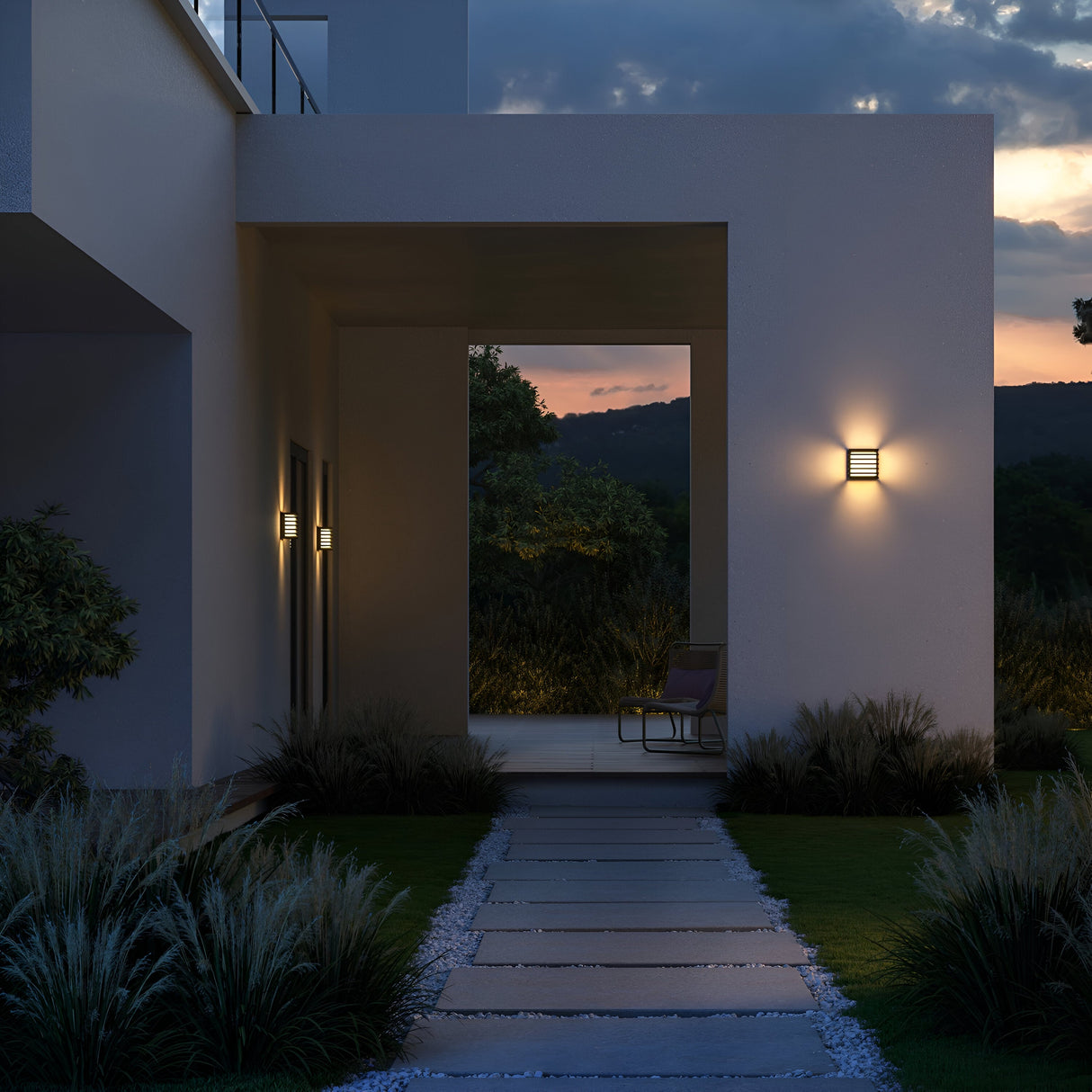 A dusk-lit pathway with grass and shrubs, illuminated by the energy-efficient Downing Street Outdoor LED Wall Light - Black (O020WL-L10B4K). The mix of cloudy and clear sky, with hints of sunset colors, enhances these IP65 outdoor fixtures leading to a seating area.