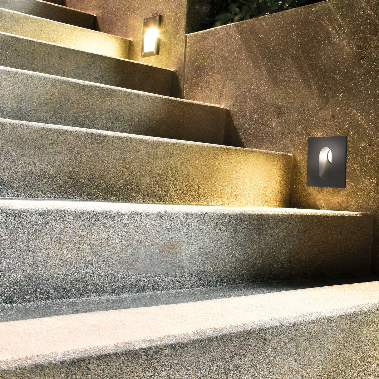 A dimly lit set of concrete stairs is softly illuminated by Via Urbana Outdoor LED Recessed Wall Lights, 4000K, 3W, Grey. With an IP54 rating and corrosion-resistant design, these lights create a warm ambiance that highlights the textured surfaces of the steps and walls.