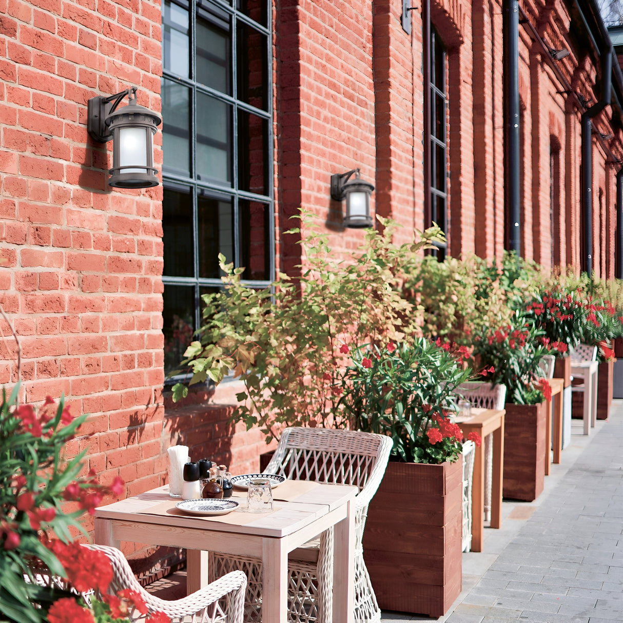 An outdoor café features wicker chairs and a wooden table against a brick building, adorned with plants and retro decor. Large windows and Salamanca Outdoor Wall Lights in bronze create a cozy, inviting atmosphere.