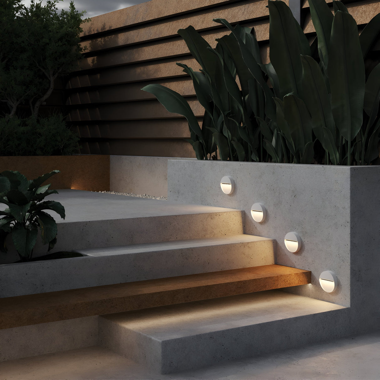 Concrete steps featuring the Mane Outdoor LED Round Wall Light - White guide you to a terrace adorned with large green plants and a wooden privacy screen. The energy-efficient LED lighting softly enhances the space, creating a modern, serene ambiance that embodies contemporary aesthetics.
