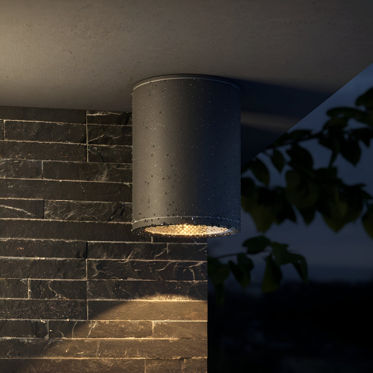 The Bar Outdoor LED Downlight 7W 3000K - Graphite, with an IP65 rating, is mounted on a damp stone wall. Its cylindrical shape casts a warm glow that illuminates the surface below with leaf silhouettes in the background.