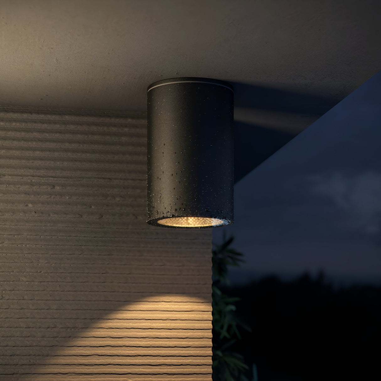 At dusk, the Roll Outdoor Downlight - Black is mounted on a textured wall, casting light over a dark background with foliage. The durable aluminum fixture has an IP54 rating and is adorned with water droplets.