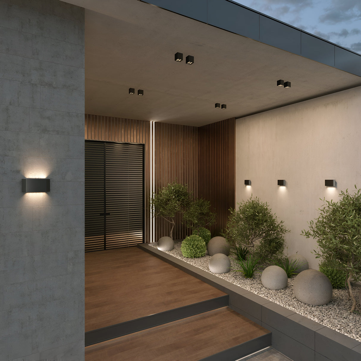 A modern entrance showcases a wooden and concrete design, with steps leading to a door with horizontal slats. Potted plants and smooth stones on a gravel bed are illuminated by the Ares Outdoor LED Square Downlight in Graphite, enhancing the evening ambiance beneath a cloudy sky.