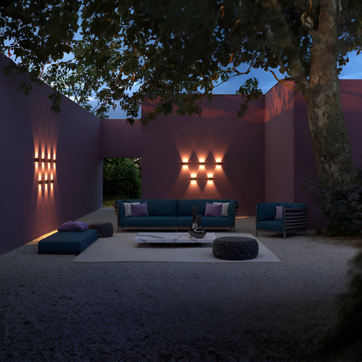 An outdoor seating area features blue sofas, a striped chair, and a low table on gravel. Purple walls, enhanced by Strato Outdoor LED Wall Lights (4W 3000K - Black), form an open courtyard. A large tree provides partial shade, creating a cozy enclosed atmosphere.