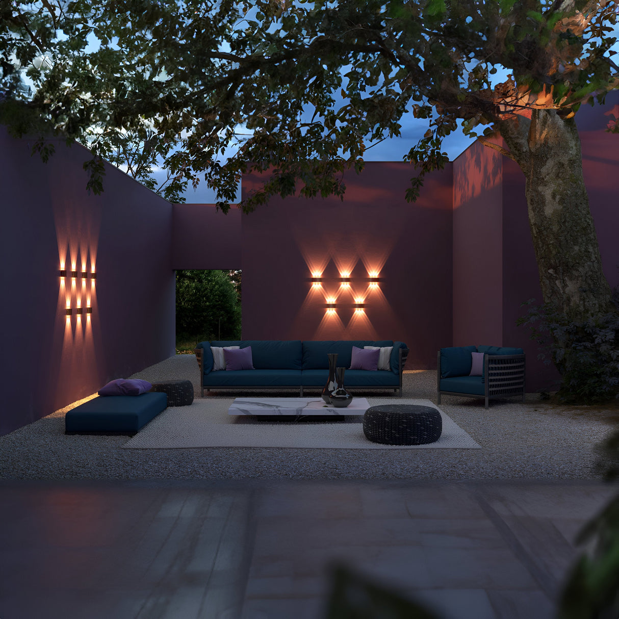A modern outdoor lounge with purple walls features teal sofas, a coffee table, and the Strato Outdoor LED Wall Light 4W in Grey. A gravel floor adds texture, while greenery peeks through an opening in the back wall, enhancing the serene ambiance.