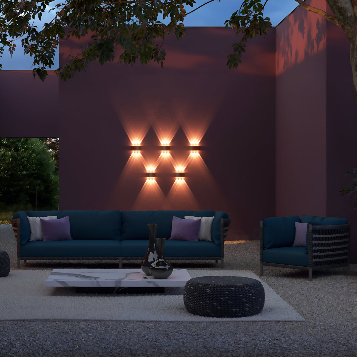 The outdoor seating area showcases a dark teal sofa and chair on a gravel patio. A low marble coffee table and round ottoman add to the elegance. The Strato Outdoor LED Wall Light, 6W 3000K in Black, casts a warm glow while tall trees lend dappled shadows to the space.