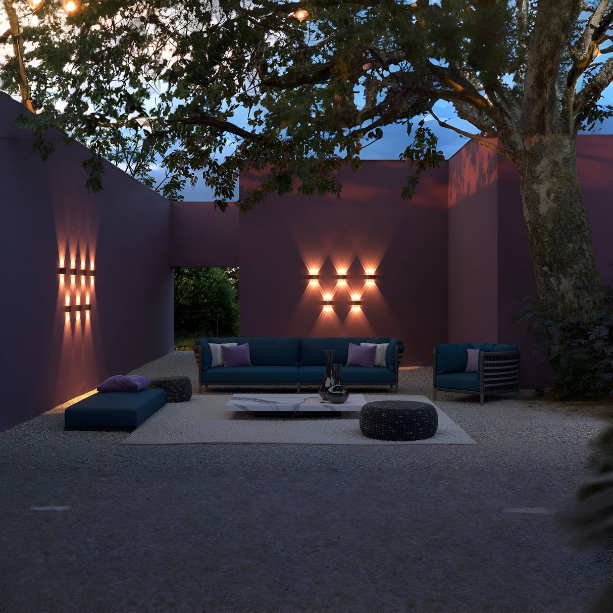 The outdoor lounge, framed by purple walls and Strato Outdoor LED Wall Lights (6W 3000K - Grey), radiates a warm glow. Blue seating surrounds a low white table, shaded by a large tree, as the IP54-rated fixtures stylishly illuminate the space with corrosion-resistant elegance at dusk.
