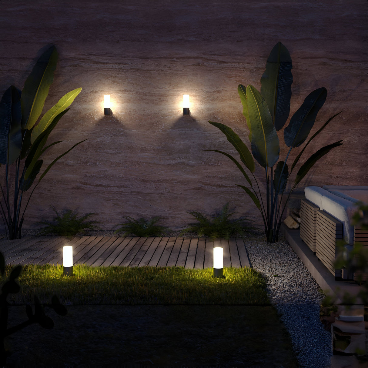 An outdoor patio features two IP54-rated wall lights illuminating a stone wall with leafy plants. A wooden deck with Willis Bollard Light 30cm in grey is bordered by grass and gravel, while an elegant seating area is partially visible on the right.