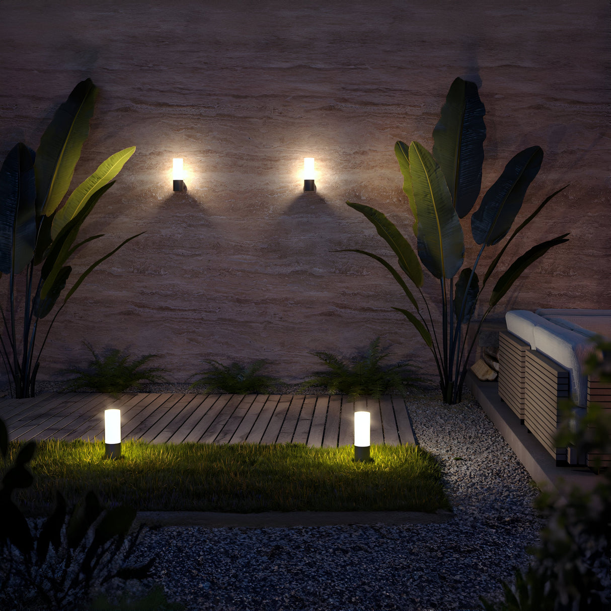 The nighttime patio has a wooden deck with two path lights on grass surrounded by gravel. Three Willis Outdoor Wall Lights in graphite (IP54-rated) cast a warm glow on the textured wall framed by tall plants with broad leaves.