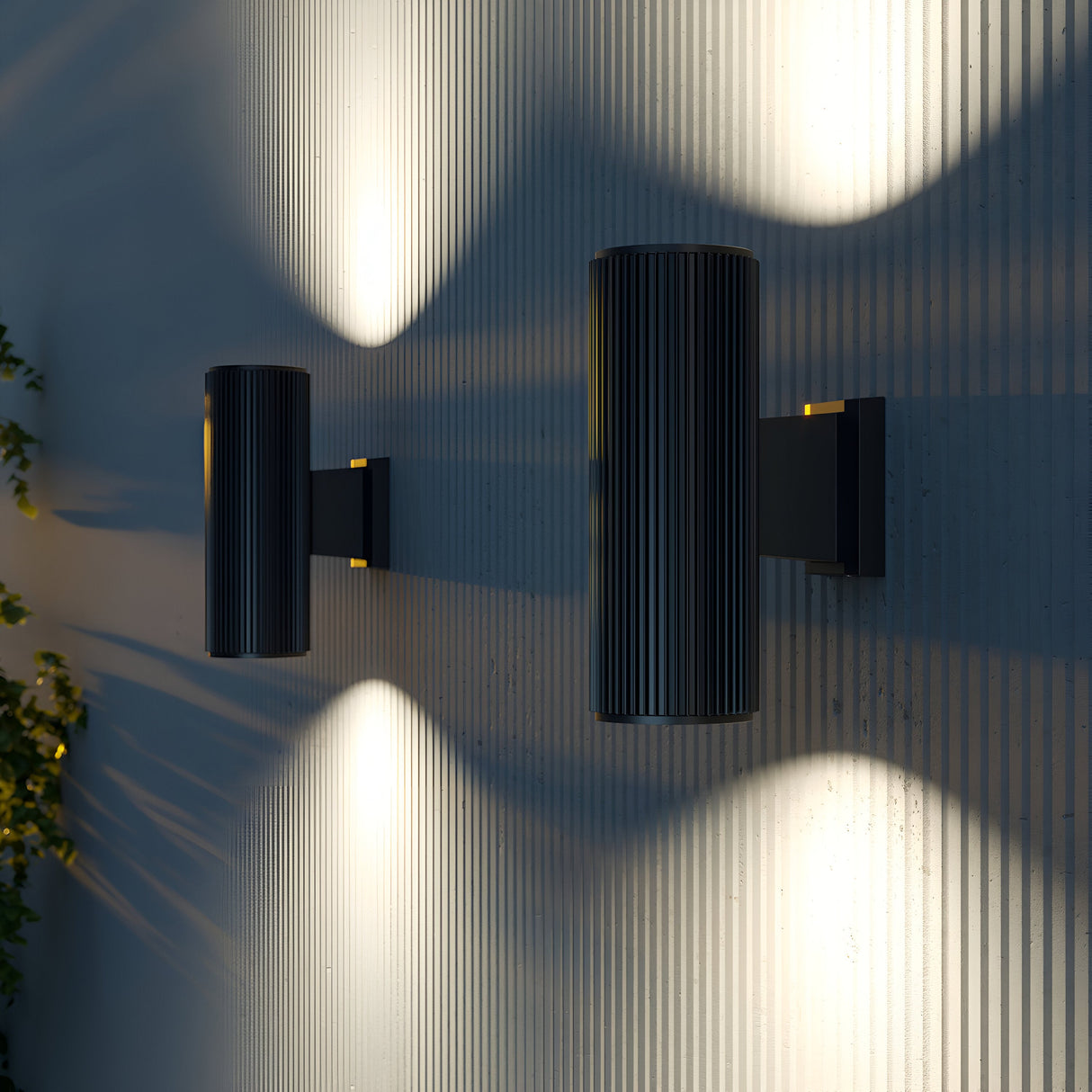 Mounted on a textured surface, two Rando Outdoor Up & Down Wall Lights in matte black emit soft yellow beams, casting an up-and-down lighting pattern. A hint of greenery on the left enhances the ambiance.