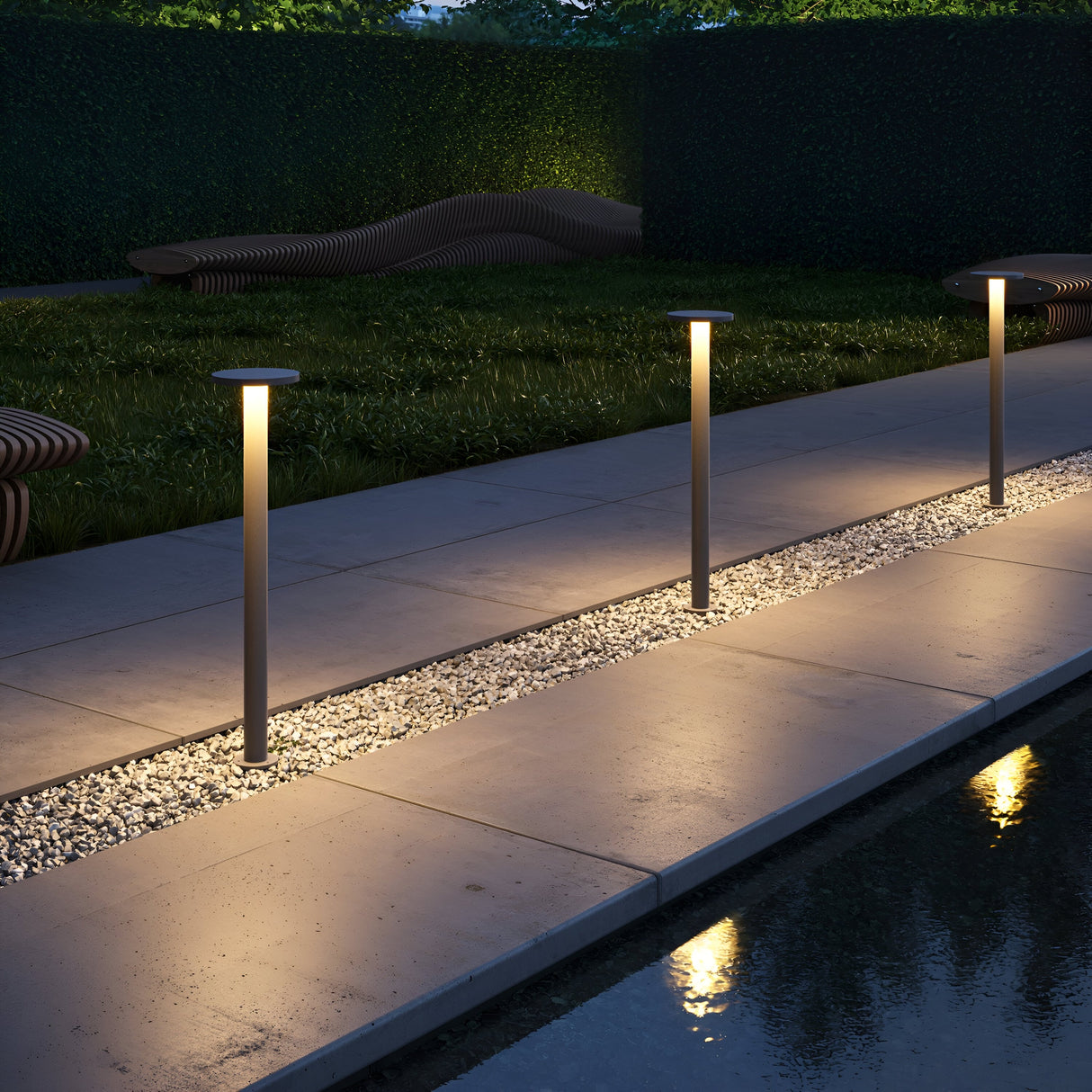 A modern outdoor pathway is illuminated by Wald LED Bollard Light 3000K 12W - Graphite, 80cm, showcasing a stone walkway lined with white pebbles. The lush grass and a sculptural wooden bench enrich the serene dusk ambiance, with the lightings IP65 rating ensuring durability.