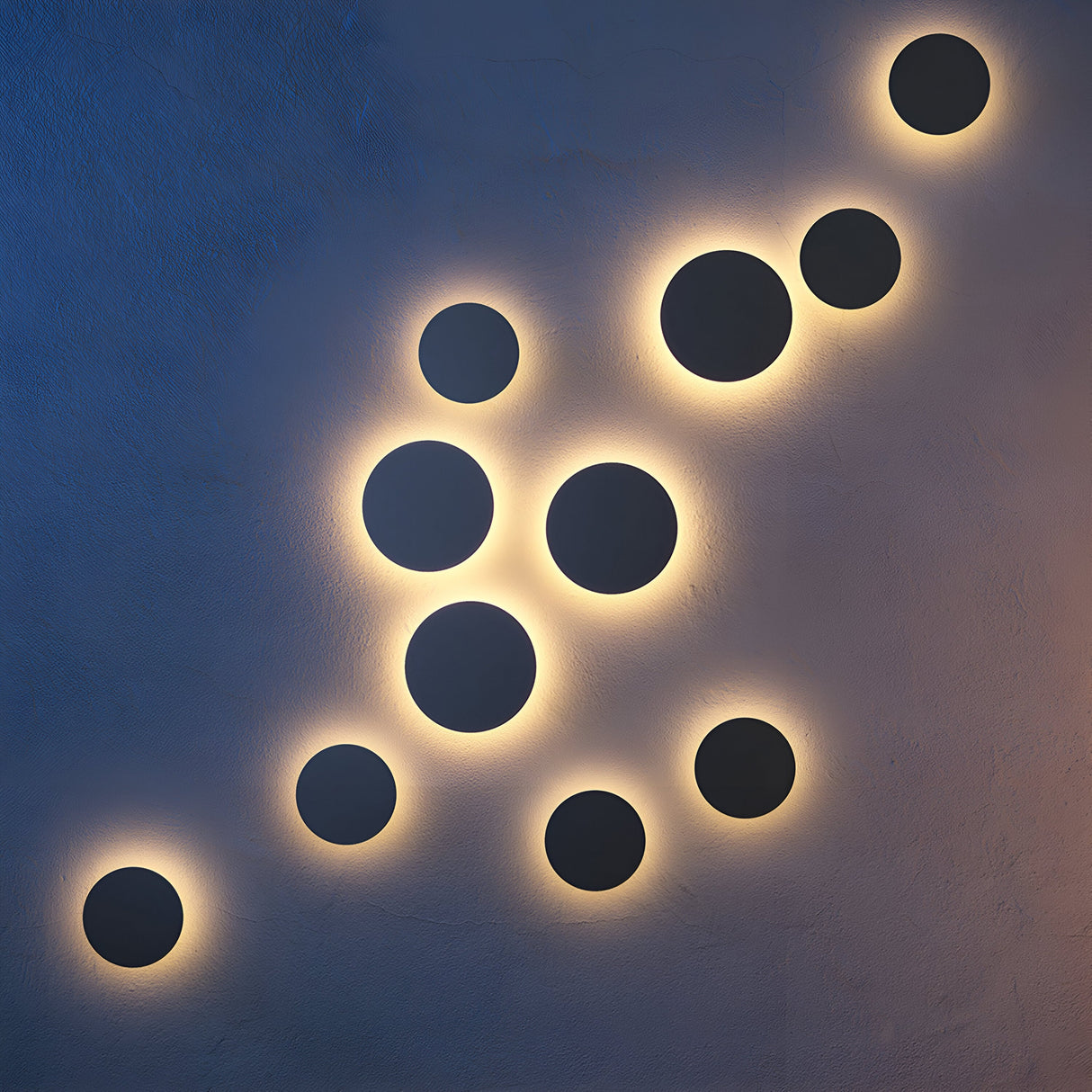 The Wald Outdoor LED Wall Light 12W 3000K - Graphite has glowing circular lights in a scattered diagonal pattern. Each IP65-rated, corrosion-resistant circle emits a warm halo against the dark wall, perfect for any outdoor setting.