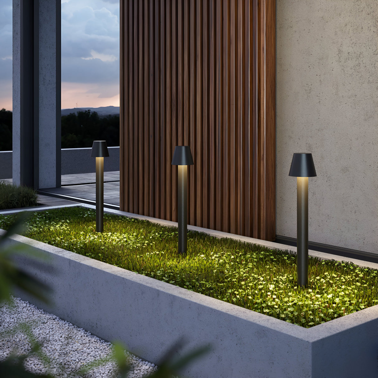 A modern outdoor area at dusk features a concrete planter box with green grass, illuminated by three sleek Harz LED Bollard Lights 80cm in graphite. A wooden slatted wall is on the right, with a cloudy sky adding drama to the scene.