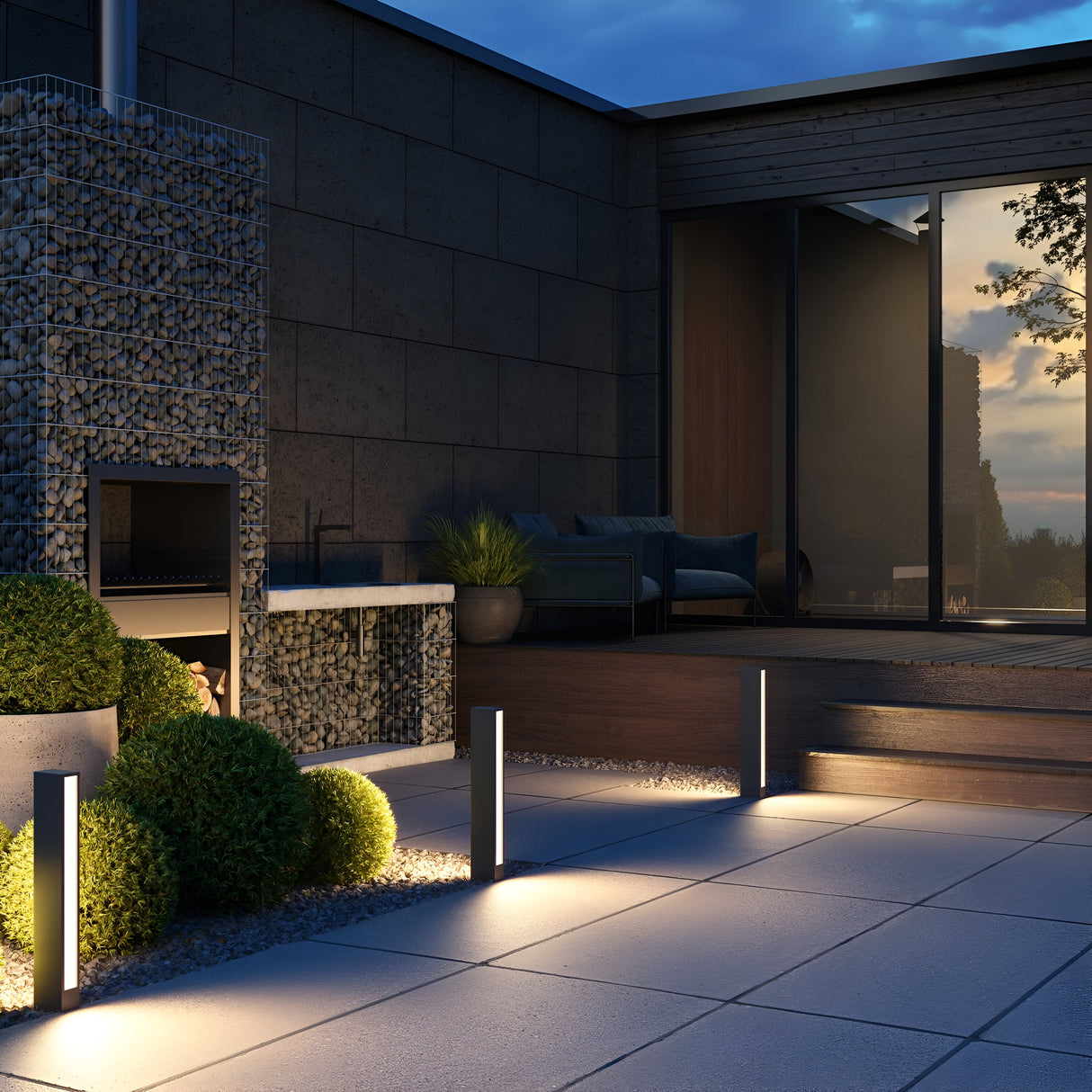A modern outdoor patio at dusk showcases a pebble-filled wall with a built-in fireplace. Strategically placed Hof LED Pedestal Lights 60cm in Graphite illuminate the pathway and neatly trimmed bushes, while a glass door reflects the evening sky, leading to the interior.