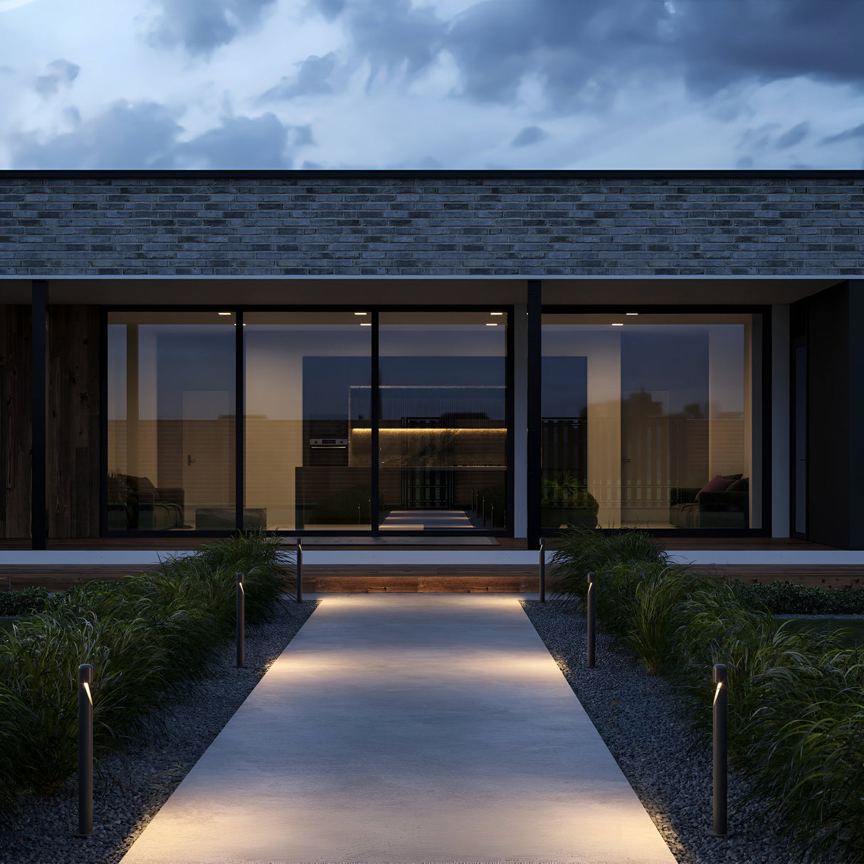 A modern house exterior at dusk features expansive glass windows. A well-lit concrete path with Hagen LED Bollard Light 60cm in graphite guides visitors to the entrance, bordered by grassy plants, while clouds create a serene backdrop.