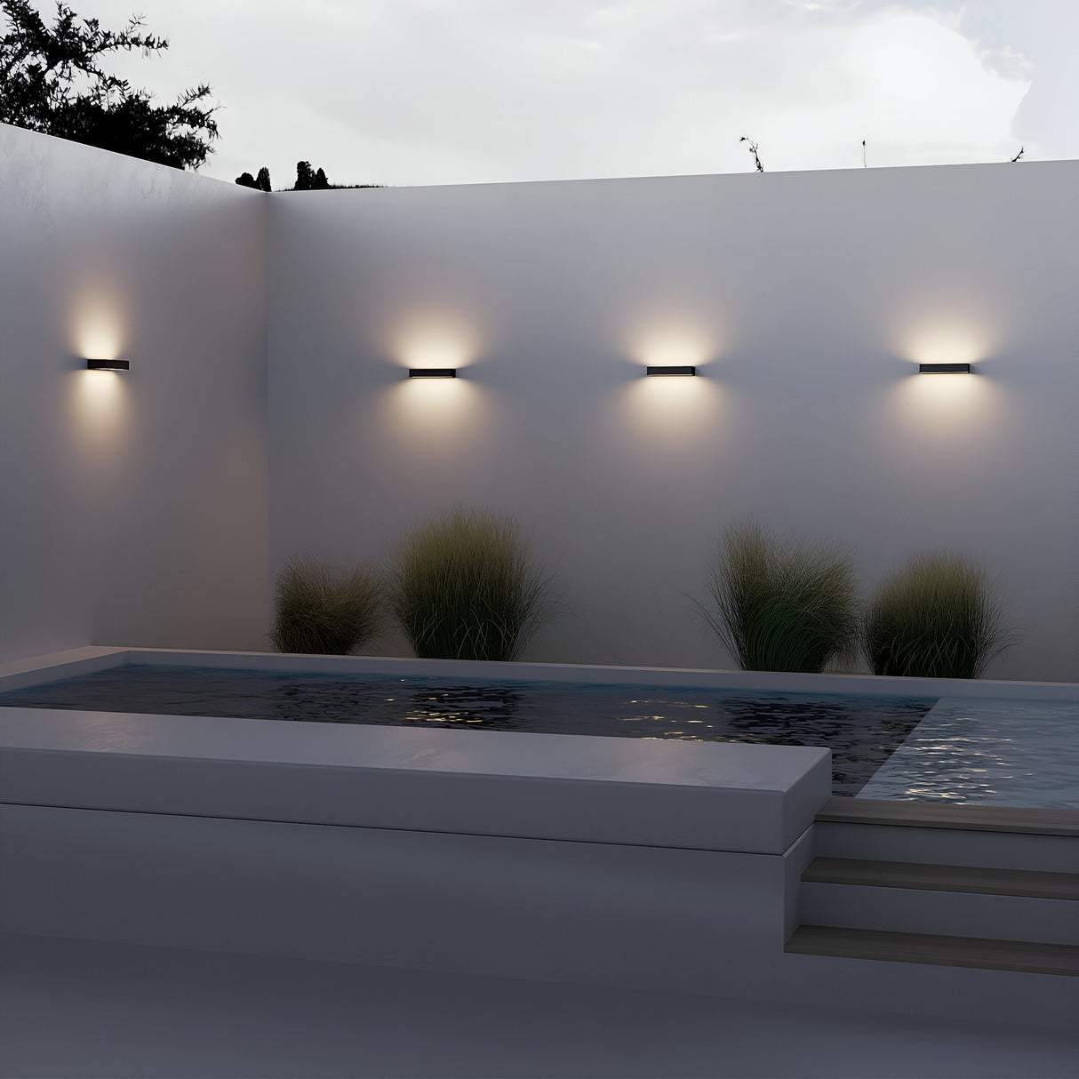 A modern outdoor pool area boasts a sleek rectangular pool, surrounded by white walls with a graphite finish. Energy-efficient Lot Outdoor LED Wall Lights softly illuminate the space, while ornamental grasses provide greenery. The overcast sky lends a serene ambiance.