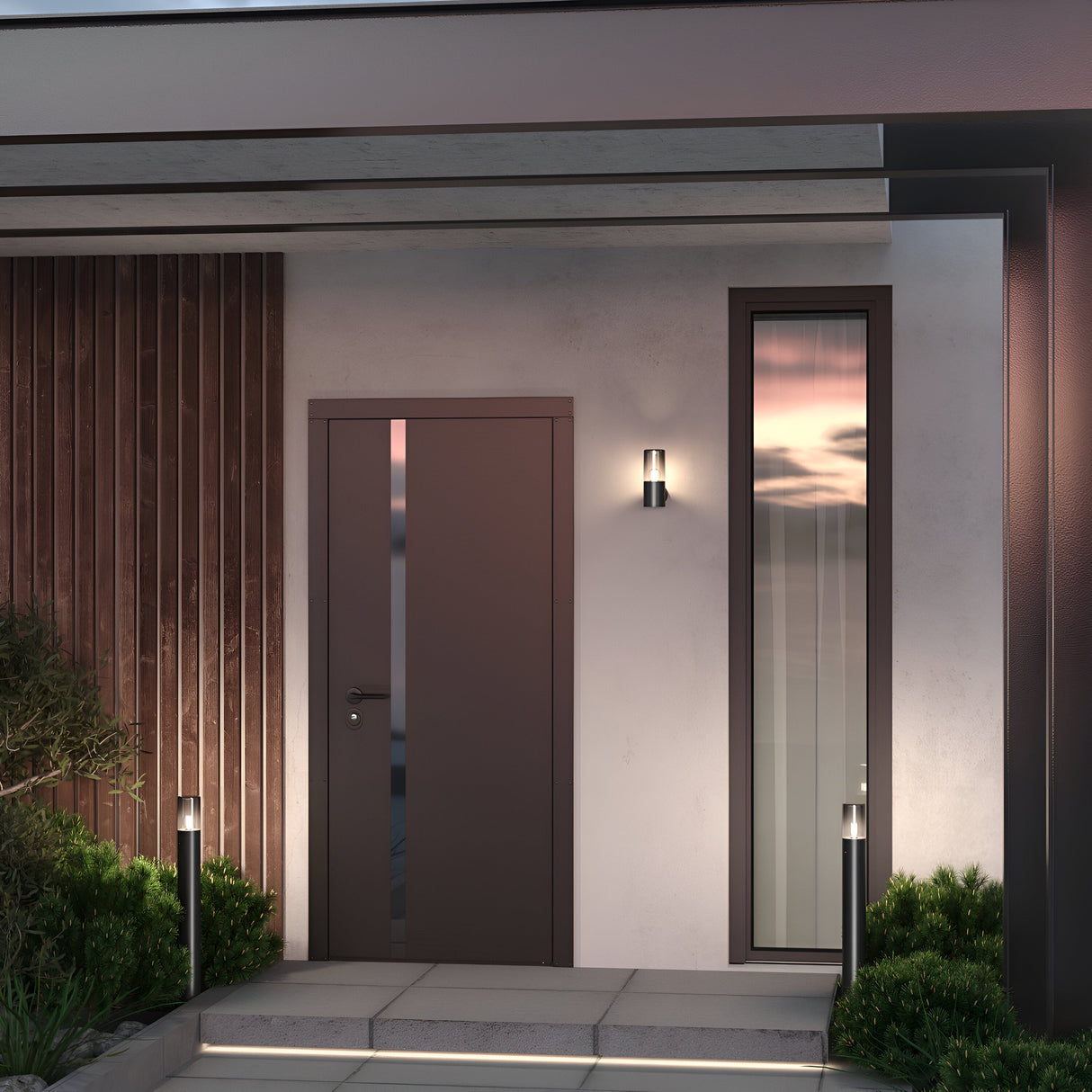 A modern house entrance features a sleek brown door with a tall, narrow window. The minimalist facade includes a wooden accent wall and soft ambient lighting. Small shrubs line the path, enhanced by Amas Bollard Light 25cm in Graphite, subtly guiding you to the door.