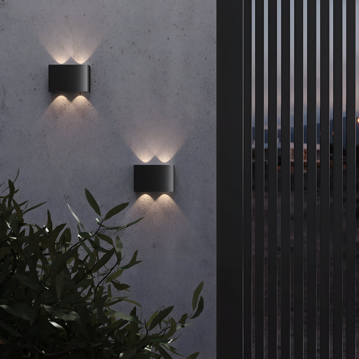 A modern exterior wall features two Show Outdoor Wall Lights (3000K - Graphite) casting sleek energy-efficient LED beams up and down. An aluminum fence stands on the right with foliage on the bottom left, all under a dimly lit sky—ideal for refined outdoor lighting at dusk.