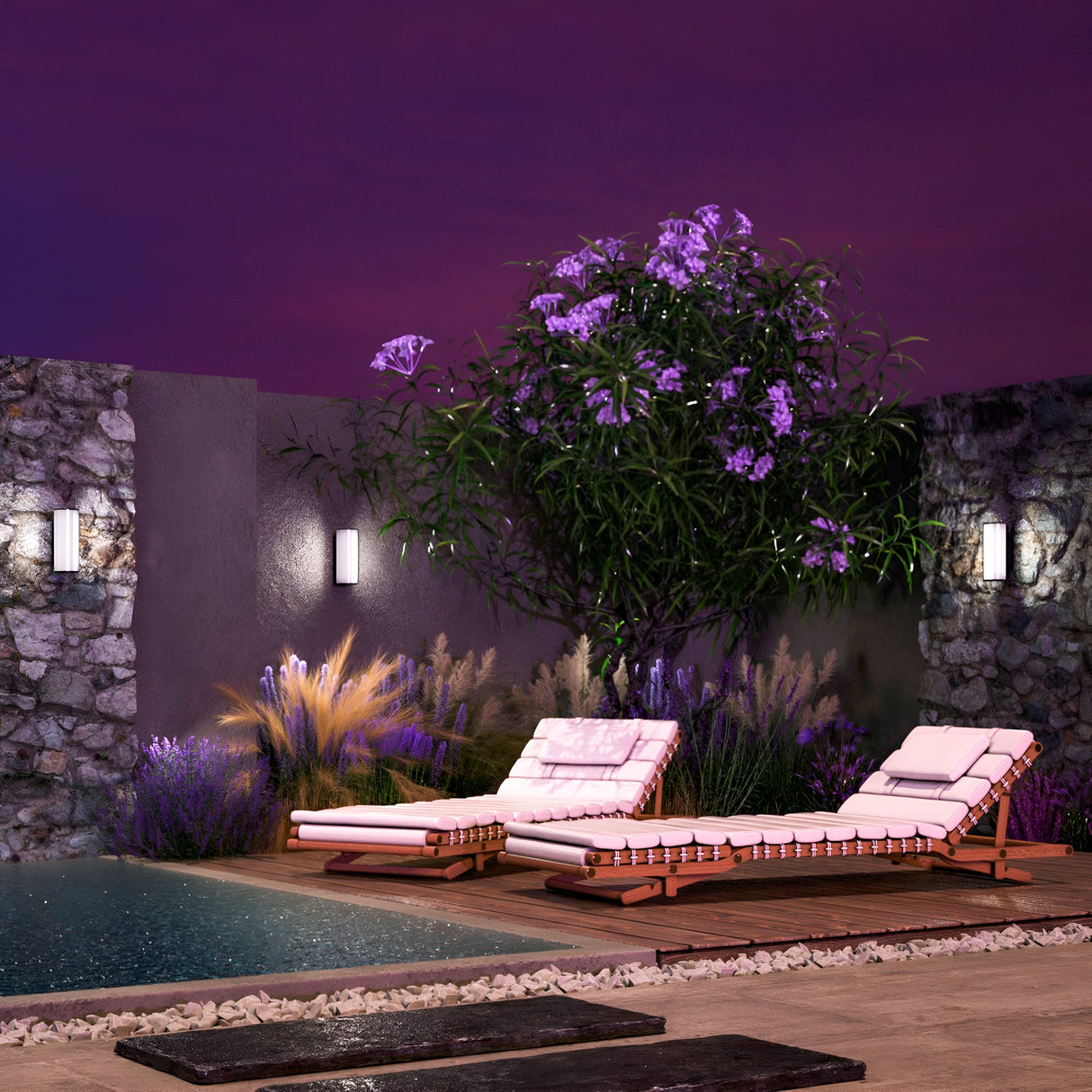 A serene backyard showcases a pair of cushioned lounge chairs on a wooden deck by a pool, adorned with purple flowers and lit by energy-efficient Rock Outdoor LED Wall Lights 3000K in Graphite against a stone wall, under a twilight sky.