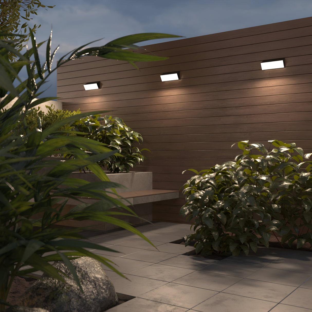 A modern patio at dusk is warmly lit by the energy-efficient Brick Outdoor LED Wall Lights in graphite, illuminating wooden paneling. Lush green plants and smooth stone tiles enhance the tranquil atmosphere, with a cozy ambiance created by the sleek graphite finish of these lights.