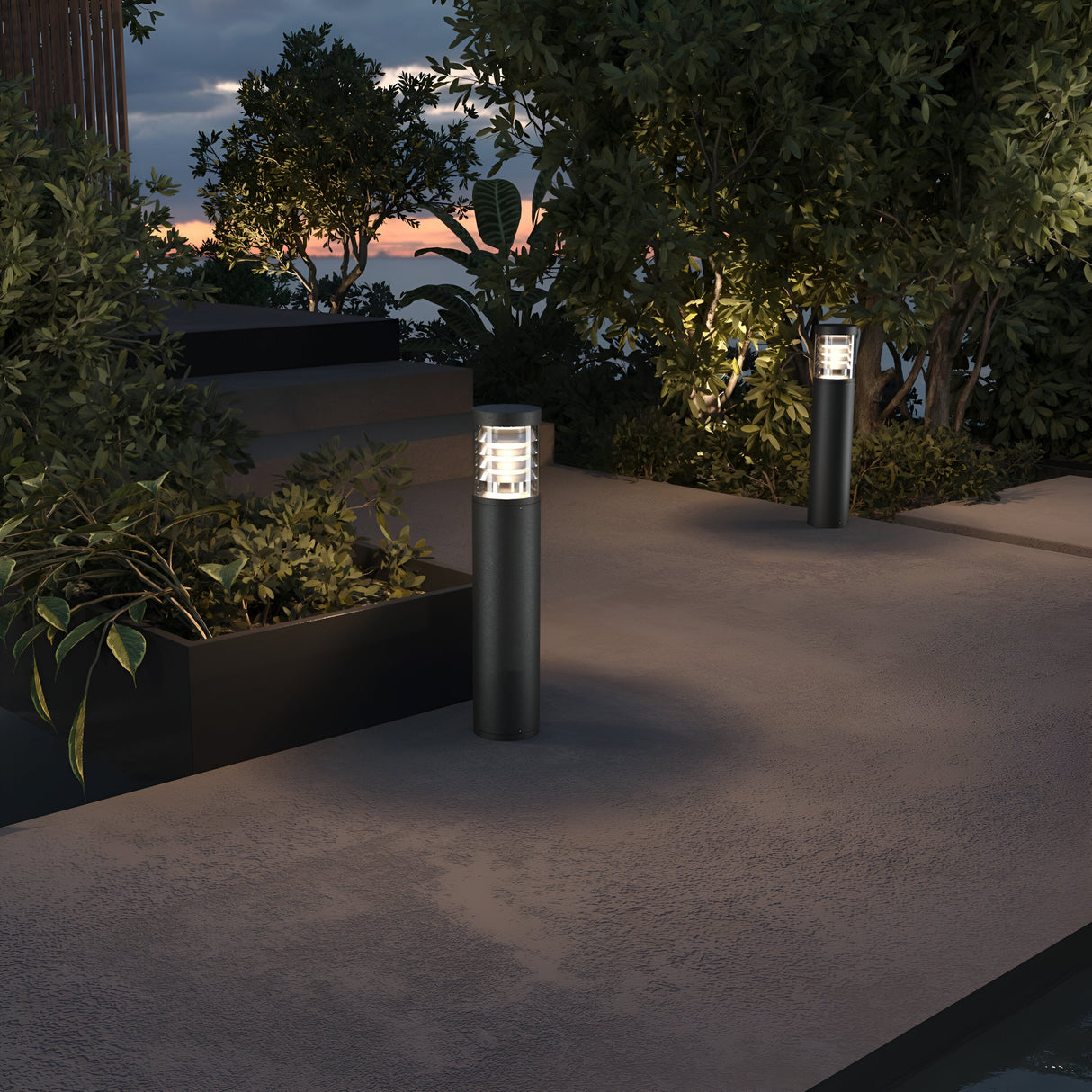 The outdoor path at dusk is illuminated by Spir Bollard Light 3000K 12W - Graphite, guiding you with its modern cylindrical glow. Surrounded by lush greenery and sunset hues, shadows dance across the textured ground from these elegantly crafted lighting fixtures.