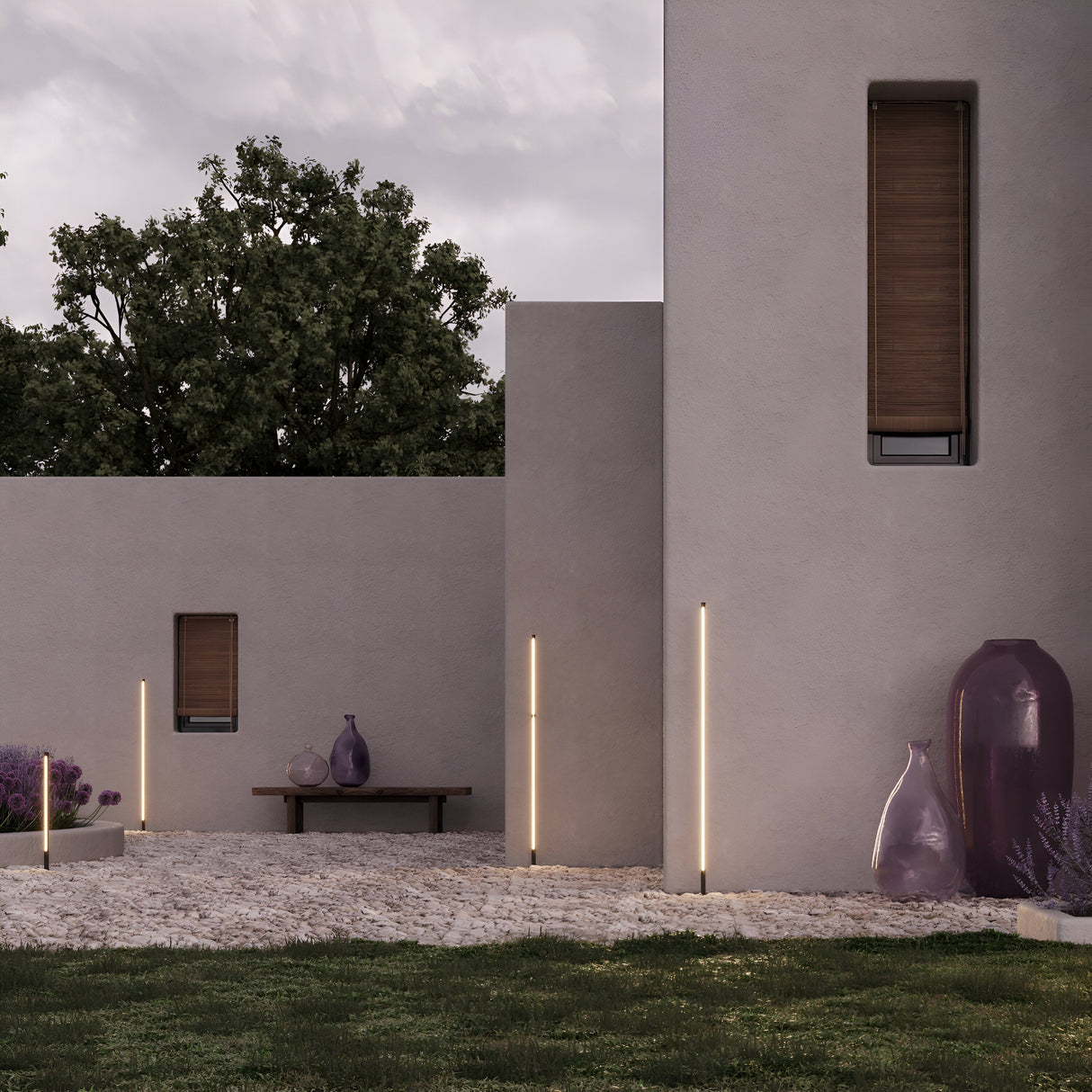 A minimalist outdoor patio with beige walls features vertical LED lights. Decorative purple vases are near lavender plants, and a wooden bench sits on a gravel path illuminated by subtle Pole LED Outdoor Spike Light 120cm - Graphite, amidst green grass and a leafy tree.